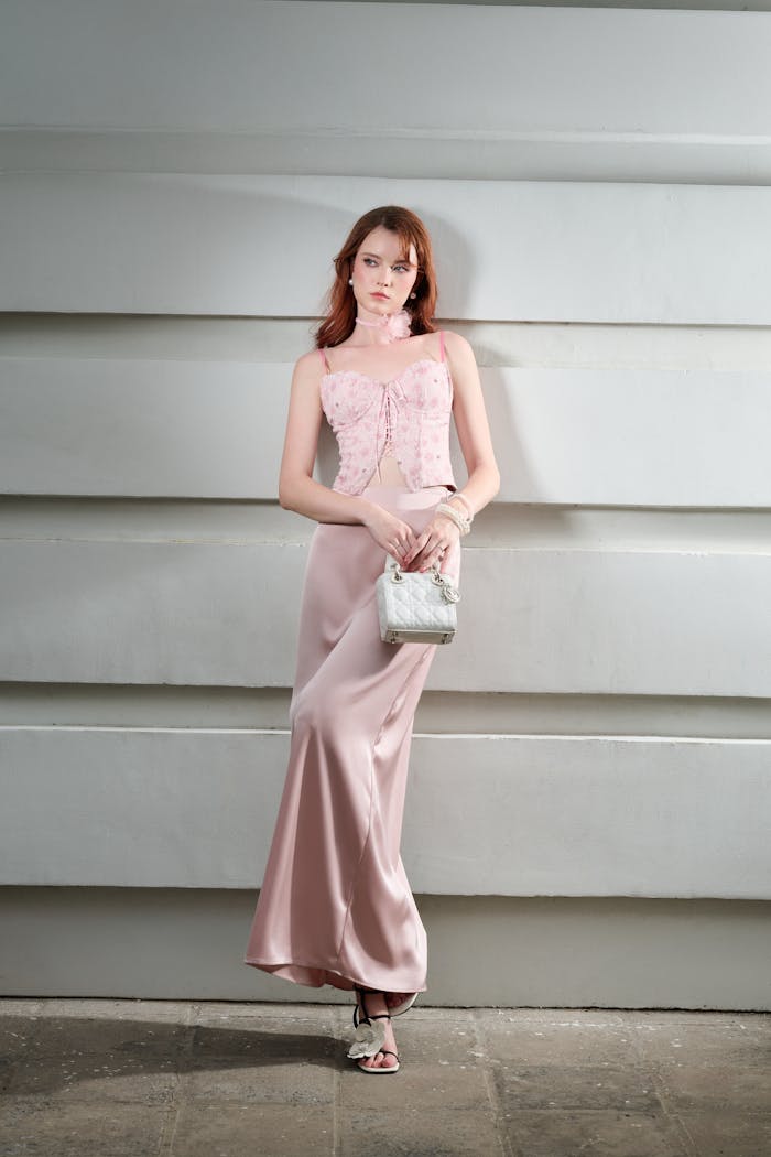 A woman in an elegant pink silk dress poses against a modern wall, exuding sophistication and style.